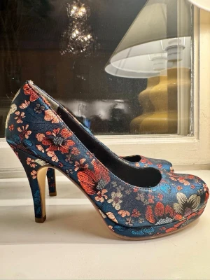 Blommiga pumps med hög klack - Snygga pumps i blått tyg med skimrande blommönster i blått, rött, lite rosa och ljusgrå. Skorna är stadiga och i hög kvalitet av Tamaris och myckwt bekväma. Hög men inte för hög klack och  elegant siluett. Perfekta för dig som vill sticka ut med ett unikt mönster och färg. Det här skorna syns! Använda två gånger och aldrig utomhus. 
