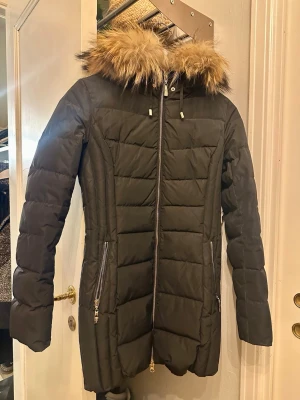 Svart lång dunjacka med äkta päls - Snygg svart lång dunjacka med dragkedja framtill och två fickor med dragkedja. Jackan har en huva med fluffig avknäppbar äkta päls i beige/bruna toner och quiltad design. Perfekt för kalla vinterdagar och riktigt varm.