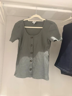Grön ribbad topp med knappar  - Snygg grön ribbad topp med korta ärmar och knappar framtill. Toppen har en djup rundringning och är i mjukt, stretchigt material som sitter skönt på kroppen. Perfekt till jeans eller kjol.