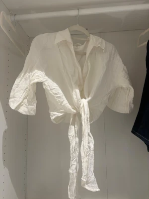 Vit knytblus med krage - Trendig vit blus med krage och knytband framtill. Blusen har trekvartsärm och är tillverkad i ett lätt, skrynkligt tyg som ger en avslappnad look. Perfekt att styla med jeans eller kjol för en cool outfit.