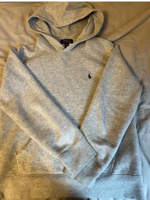 Grå hoodie från Polo Ralph Lauren - Klassisk grå hoodie från Polo Ralph Lauren med broderad marinblå logga på bröstet. Hoodien har huva, magficka och ribbade muddar vid ärmar och nederkant. Tillverkad i mjukt bomullsmaterial som är skönt att bära.
