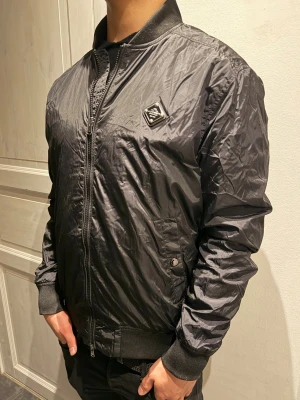 J. Lindeberg jacka  - Snygg svart bomberjacka från J. Lindeberg  med glansig finish och ribbade muddar. Jackan har dragkedja framtill, två fickor med knapp och en liten logotyp på bröstet. Perfekt för en trendig streetwear-look.