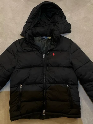 Svart pufferjacka från Polo Ralph Lauren - Svart pufferjacka från Polo Ralph Lauren med avtagbar huva och dragkedjor på fickorna. Jackan har en quiltad design och en röd logga broderad på bröstet. Perfekt för kyliga dagar och enkel att matcha med olika outfits.