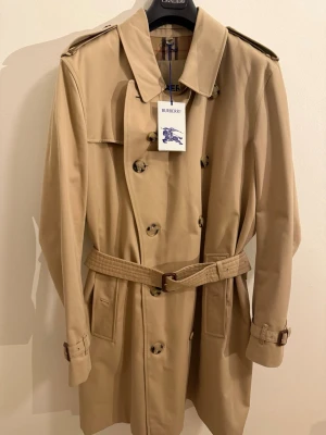 Beige trenchcoat från Burberry - Helt NY.Klassisk beige trenchcoat från Burberry, modell The Kensington. Jackan har dubbelknäppning, bälte i midjan och axelklaffar. Insidan har det ikoniska rutmönstret. Perfekt för dig som vill ha en tidlös och stilren look.