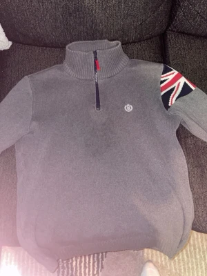 Grå stickad tröja med halvzip från Henry Lloyd - Grå stickad tröja med halvzip och hög krage. På ena axeln finns en stor Union Jack-detalj i rött, vitt och blått. Liten broderad logga på bröstet. Perfekt för dig som gillar klassisk stil med en twist.