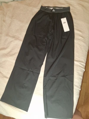 Svarta mjukisbyxor från Alo - Snygga svarta mjukisbyxor från Alo med bred resår i midjan och raka ben. Byxorna har en stilren look och är tillverkade i ett mjukt och stretchigt material som känns skönt mot huden. Perfekta för en avslappnad och trendig stil.