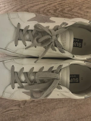 Vita sneakers från Superga med stjärna - Snygga vita sneakers från Superga med grå stjärndetalj på sidan och matchande grå skosnören. Skorna har en klassisk låg modell och svart sula undertill. Perfekta för en stilren och avslappnad look.