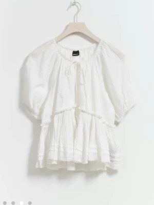 Vit volangblus med puffärm - Söt vit blus från Gina med volanger och puffiga korta ärmar. Blusen har rund halsringning med knytband och är tillverkad i ett lätt, transparent tyg som ger en luftig känsla. Perfekt för en trendig och avslappnad look.