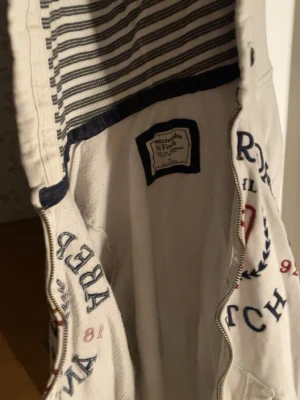 Vit hoodie från Abercrombie & Fitch - Vit hoodie med dragkedja från Abercrombie & Fitch. Stor logga och tryck framtill i marinblått och rött, samt fickor på magen. Randigt innerfoder i huvan och klassisk college-stil. Perfekt för en avslappnad och sportig look.