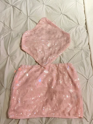 Rosa paljettset med kjol och topp - Säljer ett glittrigt tvådelat set i ljusrosa med paljetter. Setet består av en minikjol och en trekantig scarf-topp med tunna knytband. Materialet är mesh med insydda paljetter som ger ett skimrande intryck. Perfekt för dig som vill sticka ut med en trendig och söt look.