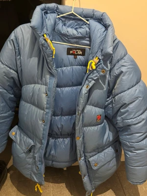Blå dunjacka Fjällräven Polar XXS - Blå dunjacka från Fjällräven Polar med gula dragskor och stora fickor framtill. Jackan har en hög krage, tryckknappar och dragkedja. Klassisk Fjällräven-logga på bröstet. Perfekt för kalla vinterdagar. Mycket använt skick