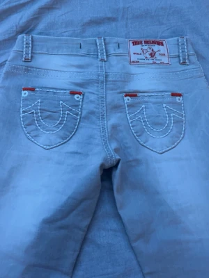 Ljusblå True Religion jeans - Säljer ett par ljusblå jeans från True Religion med ikoniska vita sömmar och detaljer i rött vid fickorna. Klassisk femficksmodell med tydlig logga på bakfickan och patch bak i midjan. Perfekta för dig som gillar streetwear och vill sticka ut. Aldrig använde helt nya nypris 1299 från vintage and friends