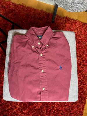 Vinröd skjorta från Ralph Lauren XS - Klassisk vinröd skjorta från Ralph Lauren med button-down krage och blå broderad logga på bröstet. Skjortan har vita knappar och är tillverkad i mjuk bomull. Perfekt för en stilren och avslappnad look.
