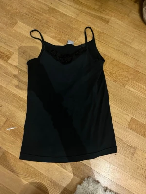 Svart linnetopp med spetsdetalj - Snygg svart linnetopp med tunna axelband och spetsdetalj vid urringningen. Perfekt att styla lager på lager eller bära som den är. Mjuk och skön känsla, enkel att matcha till olika outfits.