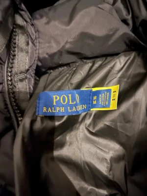 Svart pufferjacka Polo Ralph Lauren - Säljer en svart pufferjacka från Polo Ralph Lauren med klassisk röd logga på bröstet. Jackan har dragkedja framtill och en hög krage för extra värme. Materialet är glansigt och quiltat, perfekt för kalla vinterdagar.