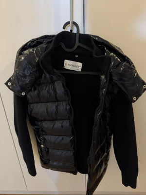 Svart dunjacka från Moncler XS - Snygg svart dunjacka från Moncler i storlek XS. Jackan har glansig dunfront och huva, samt stickade svarta ärmar och rygg. Dragkedja framtill och två sidoknappar. Perfekt för kalla vinterdagar och riktigt trendig look.