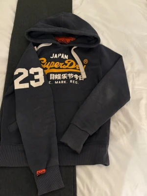  Superdry hoodie  - Snygg mörkgrå hoodie från Superdry med stort gult och vitt tryck på bröstet och ärmen. Klassisk känguruficka, vita dragsnören i huvan och ribbade muddar. Perfekt för en avslappnad och sportig stil.