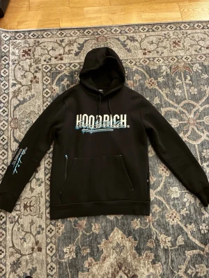 Svart hoodrich hoodie - Svart hoodie från Hoodrich med stor vit logga och blått tryck på bröstet  i vitt och blått. Klassisk huva med dragsko och känguruficka framtill. Perfekt streetwear-vibe.