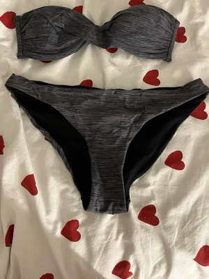 Mörkgrå bandeau bikini - Snygg mörkgrå bandeau bikini med diskret randigt mönster. Överdelen är axelbandslös och har twistad front, underdelen är klassisk med låg midja. Perfekt för stranden eller poolen.