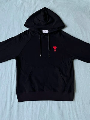 Svart hoodie från Ami Paris - Säljer en svart hoodie från Ami Paris med klassisk huva och dragsnören. På bröstet finns Ami:s ikoniska röda hjärta med A-broderi. Hoodien har känguruficka och ribbade muddar. Tillverkad i mjuk bomull för skön känsla.