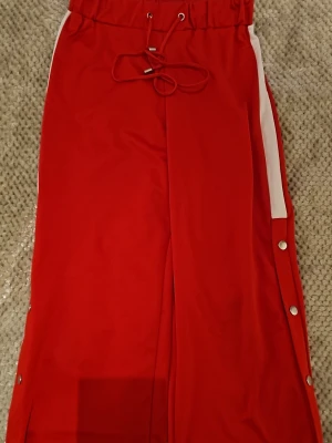 Röda mjukisbyxor Love Soul London - Snygga röda mjukisbyxor från Love Soul London med vita sidoränder och tryckknappar längs benen. Byxorna har dragsko i midjan och är perfekta för en avslappnad och sportig stil. Materialet känns mjukt och bekvämt.