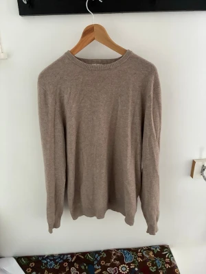 Beige stickad tröja i Kashmir  - En enkel och stilren beige stickad tröja med rund hals och långa ärmar. Tröjan har ribbade muddar vid hals, ärmslut och nederkant. Perfekt för lager på lager och passar till det mesta i garderoben. 100% Kashmir.