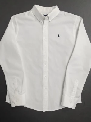 Vit Ralph Lauren Skjorta - Klassisk vit skjorta från Ralph Lauren i bomull med broderad svart logga på bröstet. Skjortan har lång ärm, knappar framtill och button-down krage. Perfekt för en clean och stilren look.