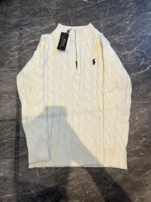 Vit kabelstickad tröja Polo Ralph Lauren - Kabelstickad vit tröja från Polo Ralph Lauren med halv dragkedja och broderad logga på bröstet. Tröjan har ribbade muddar och hög krage, perfekt för lager-på-lager. Klassisk och stilren design som funkar året runt.