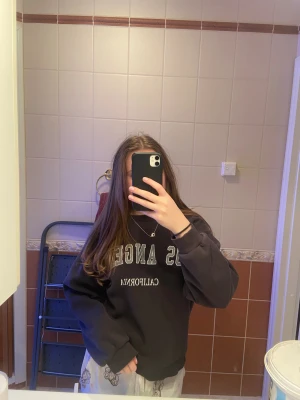Svart sweatshirt  - Cool svart sweatshirt med trycket 'Los Angeles California' i vitt och grått på bröstet. Tröjan har rund hals, ribbade muddar och en loose passform. Perfekt för dig som gillar streetstyle och vill ha en enkel men snygg look.