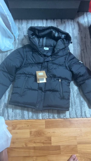  Burberry jacka - Svart pufferjacka från Burberry med huva och tydlig logotyp på ärmen. Jackan har dragkedja framtill, två fickor med dragkedja och quiltad design. Insidan av huvan har ett diskret rutmönster. Perfekt för kalla dagar och riktigt snygg streetstil.