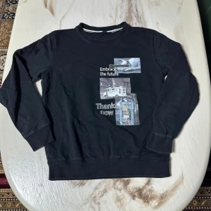 Svart sweatshirt med tryck, storlek S - Svart sweatshirt med coolt tryck på framsidan med texten 'Embrace the future' och 'Thanks now' samt bilder av katt, buss och telefonkiosk. Rund hals och långa ärmar med ribbade muddar. Perfekt för dig som gillar streetstyle och grafiska prints.