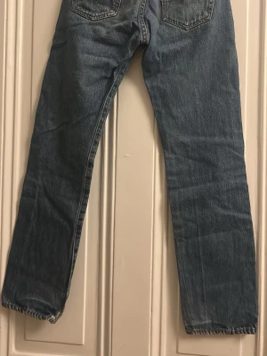 Levi's 501 raka jeans blå denim - Klassiska Levi's 501 jeans i blå denim med raka ben och normal passform. Jeansen har fem fickor, knappgylf och den ikoniska läderpatchen bak i midjan. Perfekta för en avslappnad och tidlös look.