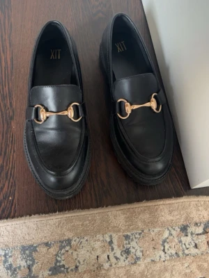 Svarta loafers med gulddetalj från XIT - Stilrena svarta loafers från XIT med chunky platt sula och snygg guldfärgad metallspänne över foten. Skorna har rund tå och är tillverkade i skinnimitation som ger en klassisk och cool vibe. Perfekta för dig som gillar en trendig och tidlös look.