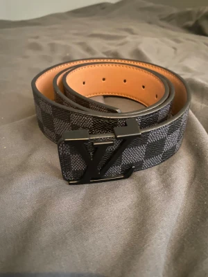 L V rutig bälte med L V-spänne - Snyggt bälte från L V med klassiskt svart L V-spänne och rutigt mönster i grått och svart. Bältet har en stilren design med slät insida i beige och flera hål för justering. Perfekt accessoar för att lyfta din outfit.