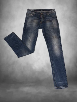 Dsqaured2 jeans - Snygga dsqaured2 jeans i klassisk blå tvätt med coola slitningar och diskreta färgstänk. Modellen har rak/slim passform och fem fickor, med detaljerade sömmar och knappgylf. Perfekt för dig som gillar en avslappnad men stilren look. Prislappar ingår vid köp
