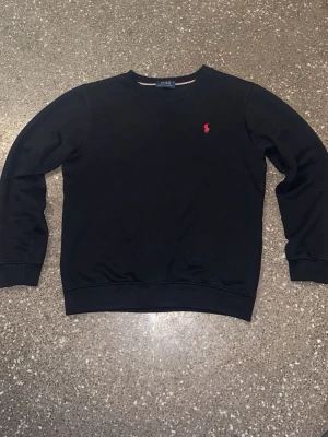 Svart tröja från Polo Ralph Lauren - Svart långärmad tröja från Polo Ralph Lauren med klassisk rund hals och rött broderat logotyp på bröstet. Tröjan är i mjuk bomull och har ribbade muddar vid ärmslut och nederkant. Perfekt för en chill och stilren look.