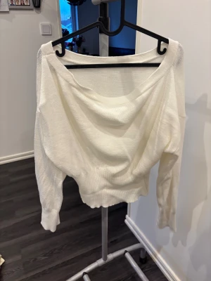 Vit offshoulder stickad tröja - Säljer en vit stickad tröja med offshoulder-design och långärmade ärmar. Tröjan har en bred halsringning som ger en avslappnad och cool look. Ribbad kant vid midja och ärmslut för extra stilpoäng. Perfekt för dig som gillar en enkel men snygg vibe.