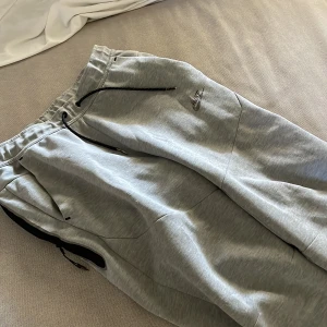 Grå mjukisbyxor från Nike - Grå mjukisbyxor från Nike med svart snörning i midjan och dragkedjeförsedda fickor på sidorna. Byxorna har en avslappnad passform och är tillverkade i mjukt bomullsmaterial. Nike-logga broderad på låret för en sportig look.