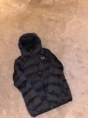 Svart pufferjacka från Under Armour - Svart pufferjacka från Under Armour med huva och två dragkedjor framtill. Jackan har ett stilrent, quiltat mönster och logga på bröstet. Materialet är glansigt och känns lätt men ändå varmt, perfekt för kalla dagar. Snygg och sportig look med normal passform.