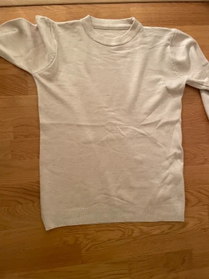Beige stickad tröja med rund hals - En enkel och stilren beige stickad tröja med rund hals och långa ärmar. Tröjan har ribbade muddar vid hals, ärmslut och nederkant. Perfekt för lager-på-lager och passar till jeans eller kjol. Mjuk och skön känsla. Helt ny utan prislapp i storlek M
