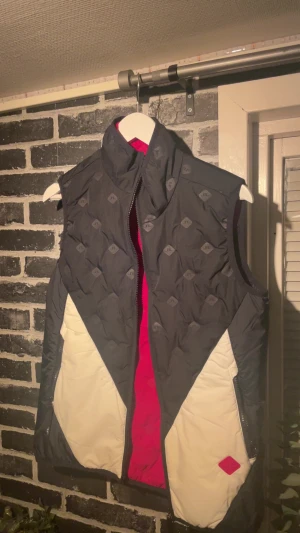 Svart och vit väst från J.Lindeberg - Snygg dunväst från J.Lindeberg i svart och vitt med röda detaljer. Västen har ett coolt mönster med JL-loggor, hög krage och dragkedja framtill. Insidan är röd och det finns praktiska fickor med dragkedja. Perfekt för lager-på-lager under kyliga dagar. Har bara användt en gång så den bara har hängt på min galje i några månader. Nu pris 3000kr kom med bud blir gärna av med den så snabbt som möjligt 