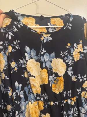 Mörkblå blommig långklänning - Supersnygg långklänning med mörkblå botten och stora gula och ljusblå blommor. Klänningen har rund hals, långa ärmar med lätt puff och resår vid ärmslut. Materialet känns mjukt och flowy, perfekt för dig som gillar romantiska vibes och blommigt mönster.