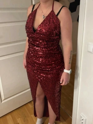 Röd glittrig långklänning med slits - Säljer en röd långklänning med glittriga paljetter, djup v-ringning och halterneck med knyt i nacken. Klänningen har en snygg slits framtill och figurnära passform som framhäver siluetten. Perfekt för dig som vill sticka ut med en glamorös look.Storlek 40/42. Köpt från bonprix