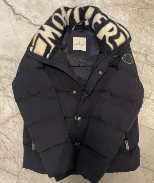 Moncler dunjacka - Snygg svart dunjacka från Moncler med stor, fluffig krage i marinblå och vit, den är exakt som den riktiga med NFC Scan, storlek M. Slitage endast på insidan men inget som är synligt för utsidan