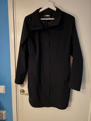 Svart regnjacka från UBR, medium - Svart regnjacka från UBR i stilren design med diskret logga vid nederkanten. Jackan har dragkedja framtill, klassisk krage och raka, långa ärmar. Perfekt för blöta dagar och har en clean look som funkar till allt.
