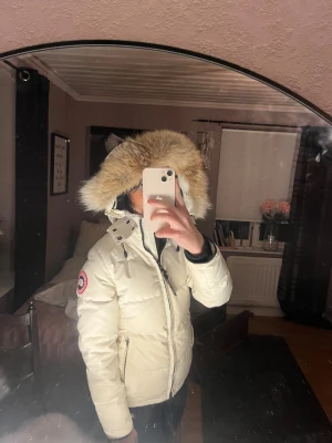 Vit dunjacka med pälskant från Canada Goose - Säljer en snygg vit dunjacka från Canada Goose med stor huva och fluffig pälskant. Jackan har dragkedja, logga på ärmen och flera fickor. Perfekt för kalla dagar och riktigt varm. Klassisk och stilren modell som funkar till allt. Äkta 