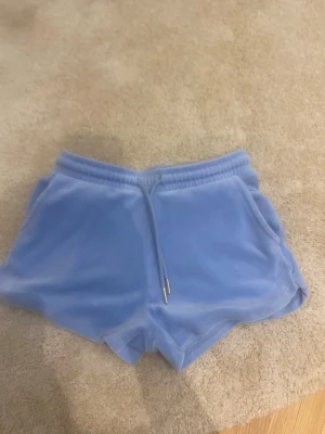 Ljusblå velourshorts Tigeri Studio - Mysiga ljusblå shorts från Tigeri Studio i mjuk velour. De har elastisk midja med snörning, fickor fram och en bakficka. Perfekta för chill dagar eller när du vill ha något bekvämt och snyggt på sommaren.