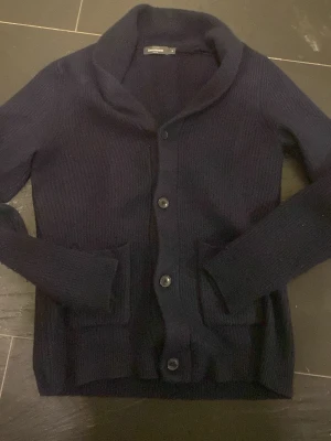 Mörkblå stickad kofta från Dressmann - Mysig mörkblå stickad kofta från Dressmann med bred krage och tre stora knappar framtill. Koftan har två praktiska fickor och långärmade ribbade muddar. Perfekt för dig som gillar klassisk och enkel stil. Annars byte mot Under armour vanish jacka stlrk L