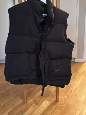 Svart dunväst från Canada Goose - Säljer en svart dunväst från Canada Goose med hög krage och två stora fickor framtill. Västen har både dragkedja och knappar, samt diskret logotyp på ena fickan. Perfekt för kalla dagar och har en clean, puffad look.