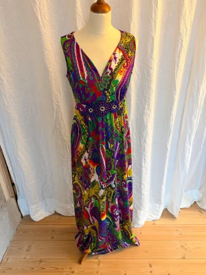 Färgstark långklänning Seppälä Woman - Säljer en maxiklänning från Seppälä Woman med färgstarkt paisleymönster i lila, grön, gul, röd och vit. Klänningen har v-ringning, ärmlös design och ett markerat midjeband dekorerat med lila pärlblommor. Perfekt statement-plagg för dig som gillar färg och unika detaljer.